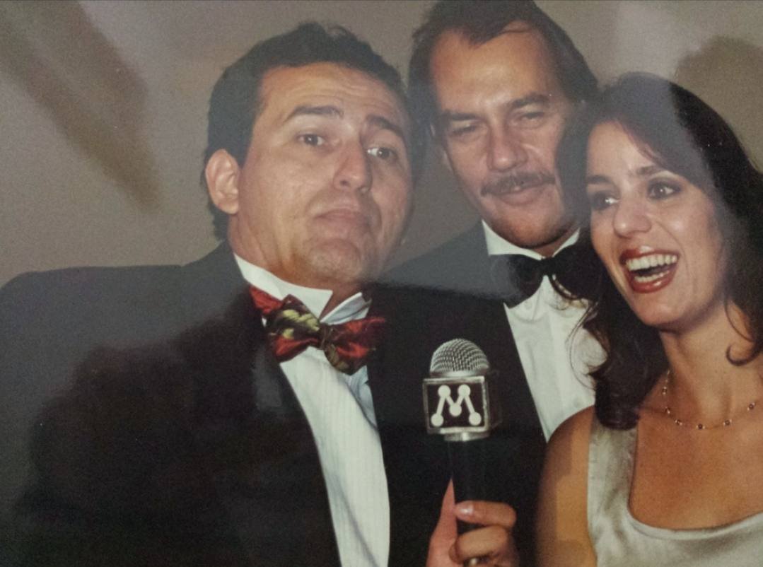 Edilásio Jr. de smoking com um casal de atores, com o microfone da TV Manchete